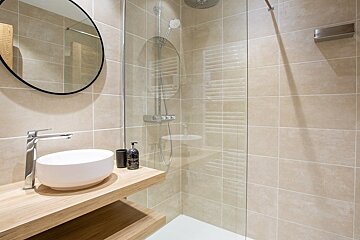 A modern bathroom with a round mirror, a white vessel sink on a wooden vanity, and a glass walk-in shower with beige tile.