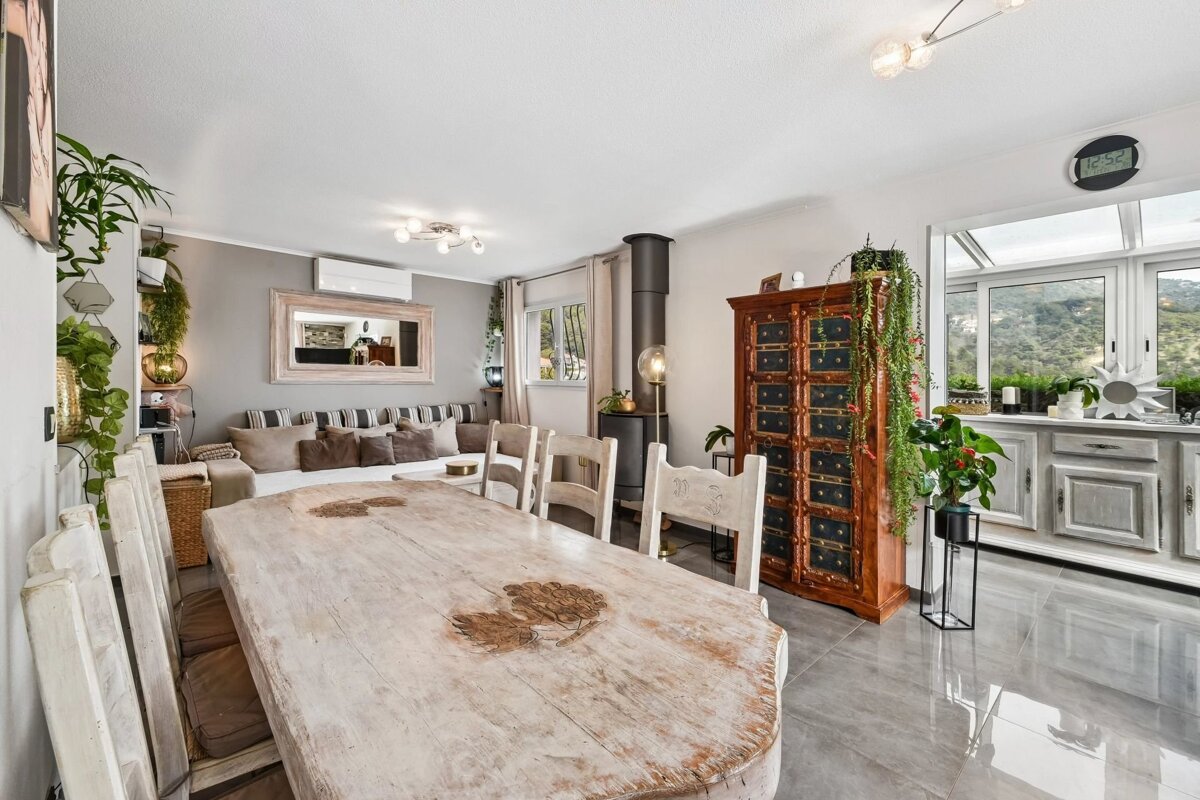 Bright, open-plan dining area with a rustic wooden table, white chairs, tiled floor, and a cozy living space in the background.