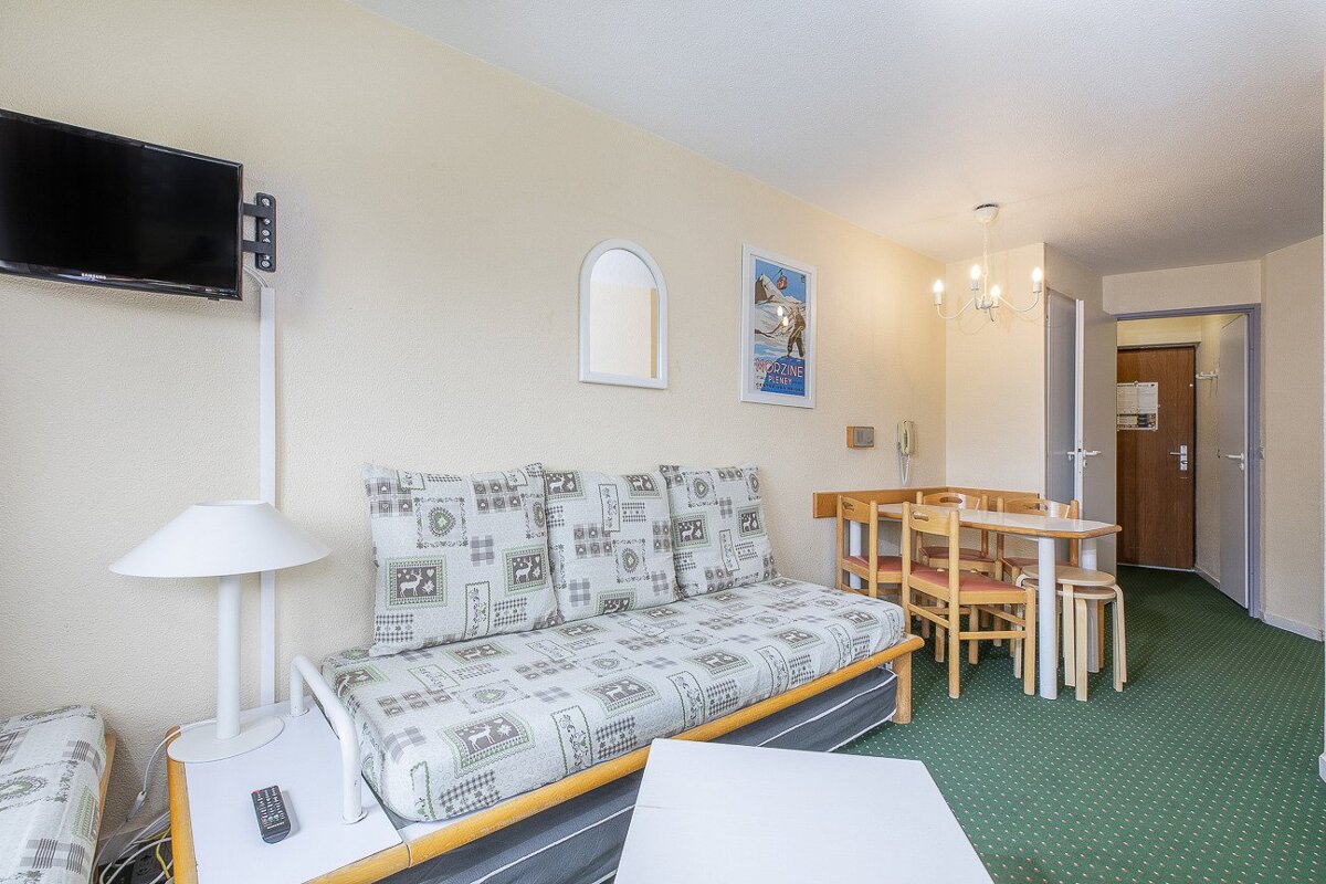 A cozy studio apartment with a patterned sofa bed, dining area, wall-mounted TV, and green carpet. Cream walls with a ski poster.