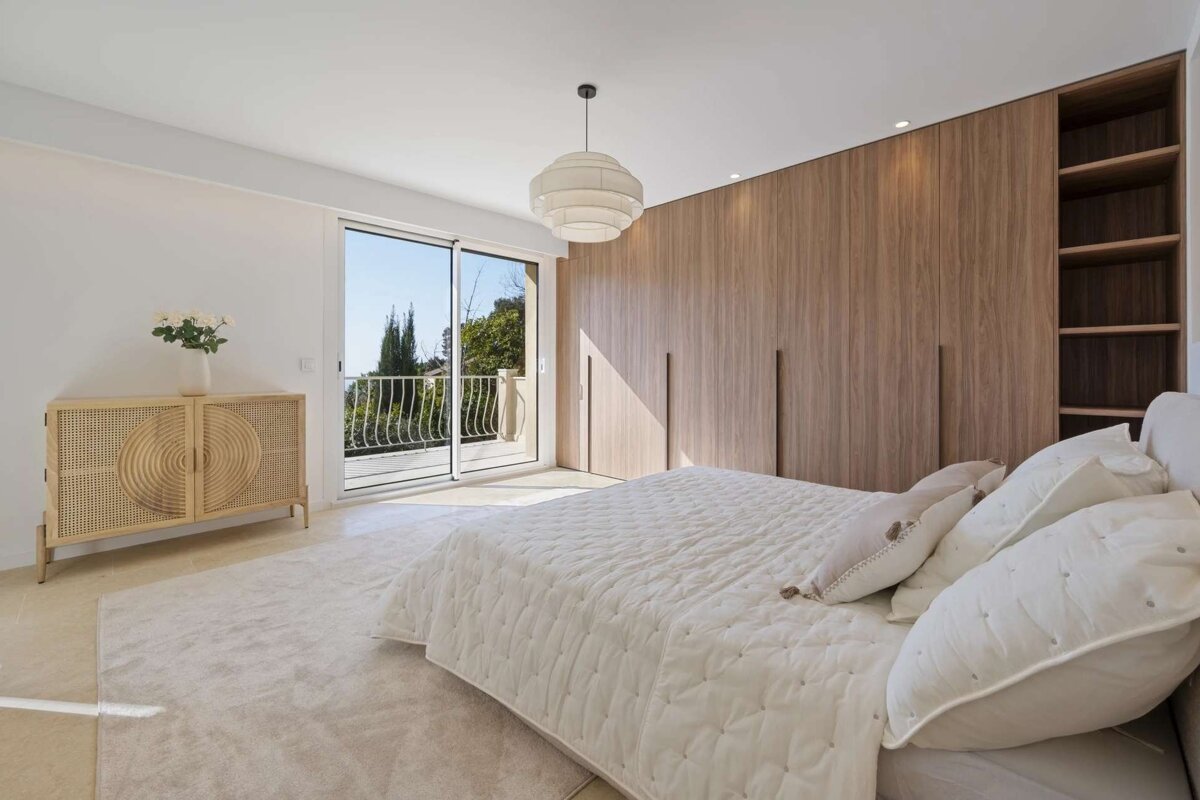 Bright, modern bedroom with a large bed, wooden wardrobe, rattan cabinet, and a balcony overlooking a scenic view.