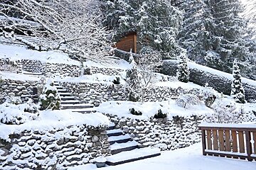 A snow-covered terraced garden with stone walls, steps, and evergreen trees, featuring a wooden cabin nestled in the snowy forest.