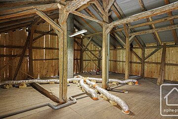 Spacious wooden attic with exposed beams, plank walls, and floor. Silver insulated ducts are laid out on the floor, under a fluorescent light.