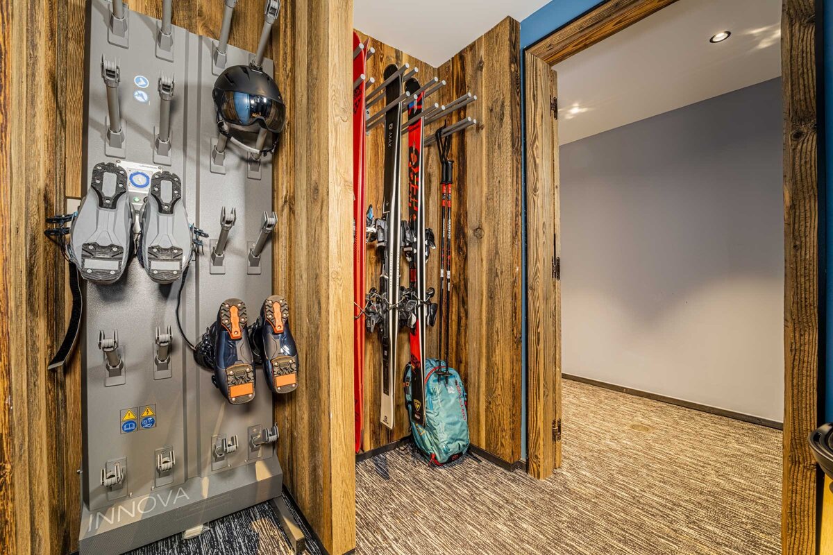 A cozy ski storage and drying area with boots, helmet, skis, and poles on racks against rustic wood paneling.