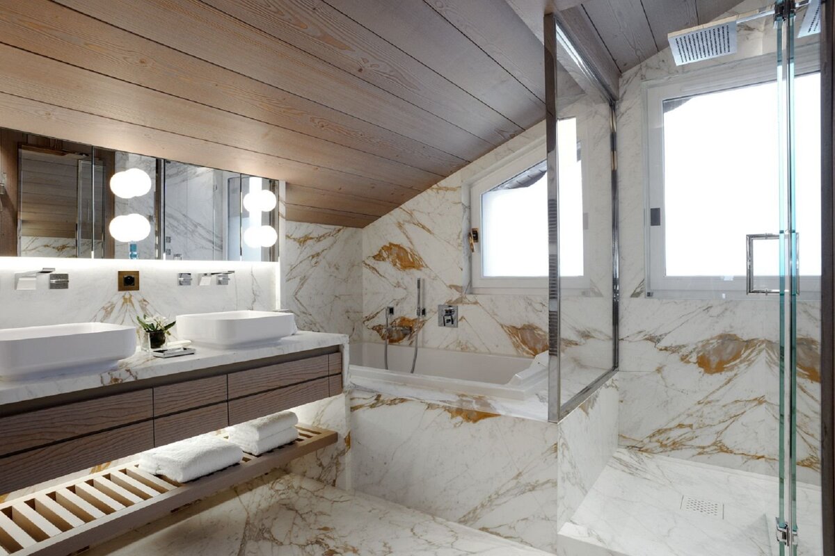 Luxurious modern bathroom with white and gold marble walls, a wooden sloped ceiling, double vanity, bathtub, and glass shower.