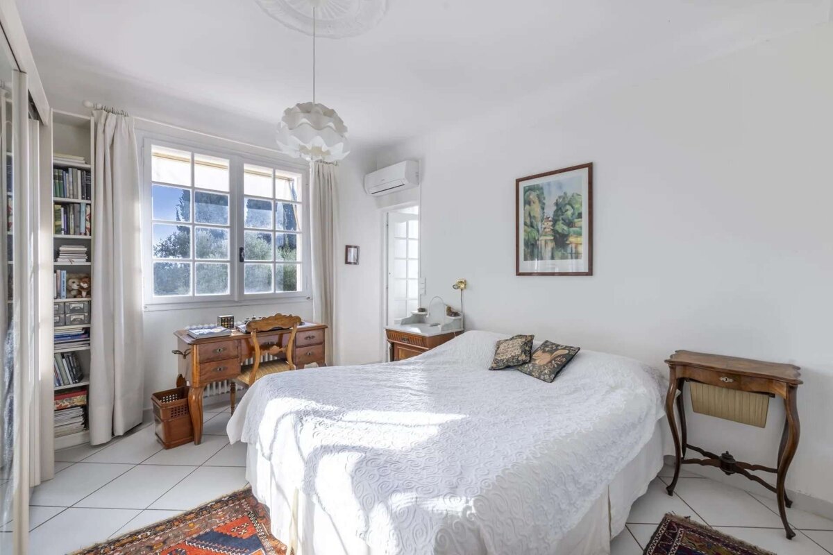 A bright, white bedroom with traditional wooden furniture, a large bed, a desk by a sunny window, a bookshelf, and tiled floor.