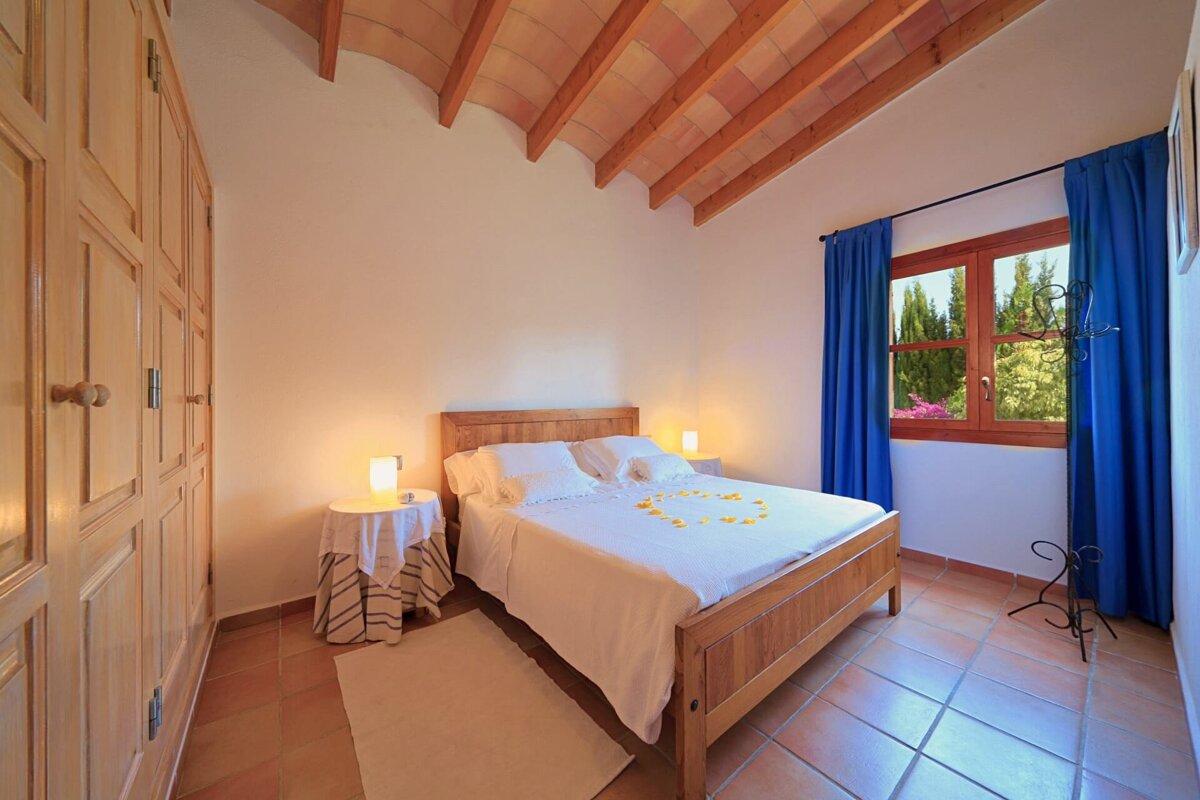 A warm, rustic bedroom features a wooden bed with white linens and petals, terracotta floor, beamed ceiling, wooden wardrobe, and a window with blue curtains overlooking greenery.