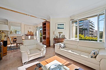 A bright living room with white leather furniture, marble floors, a dining area, and large windows offering a city view with greenery.