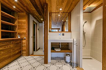A warm, modern bathroom features wood-paneled walls/ceiling, patterned floor tiles, a white vanity, large mirror, and a rain shower.