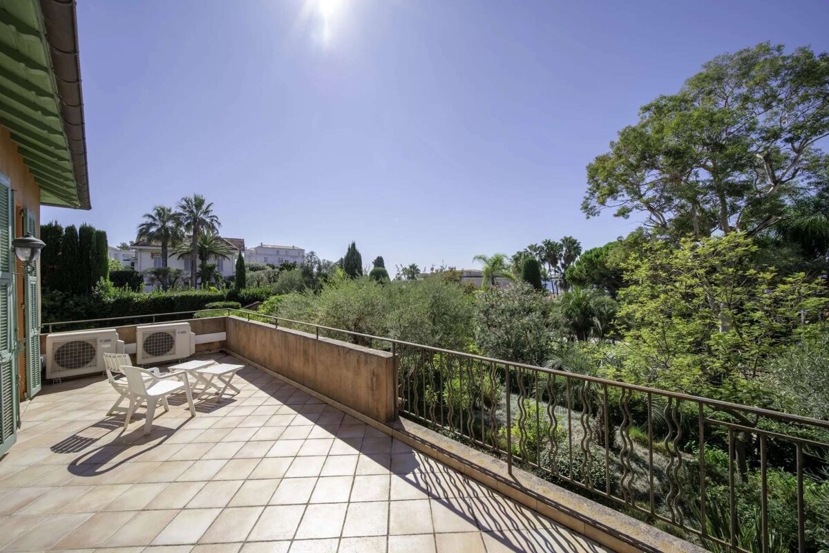 Sunny tiled balcony with chairs and AC units, overlooking a lush green garden with trees, distant houses, under a bright blue sky.