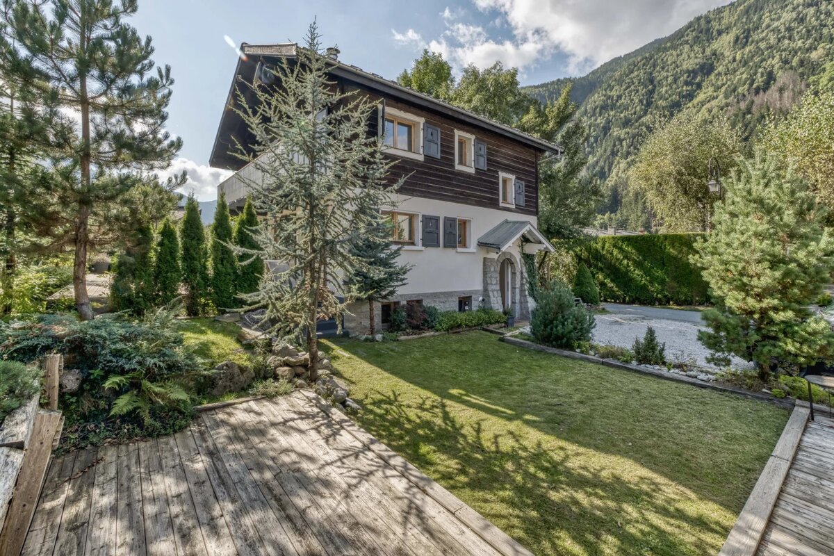 A charming chalet-style house with a lush garden and wooden deck, set against a backdrop of forested mountains under a sunny sky.