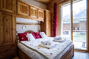 Cozy wooden cabin bedroom with red pillows, white bathrobes on the bed, and a large sliding glass door showing mountain views.