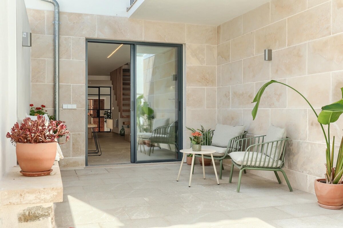 A modern patio with light stone walls, green outdoor chairs, potted plants, and a sliding glass door revealing an interior with stairs.