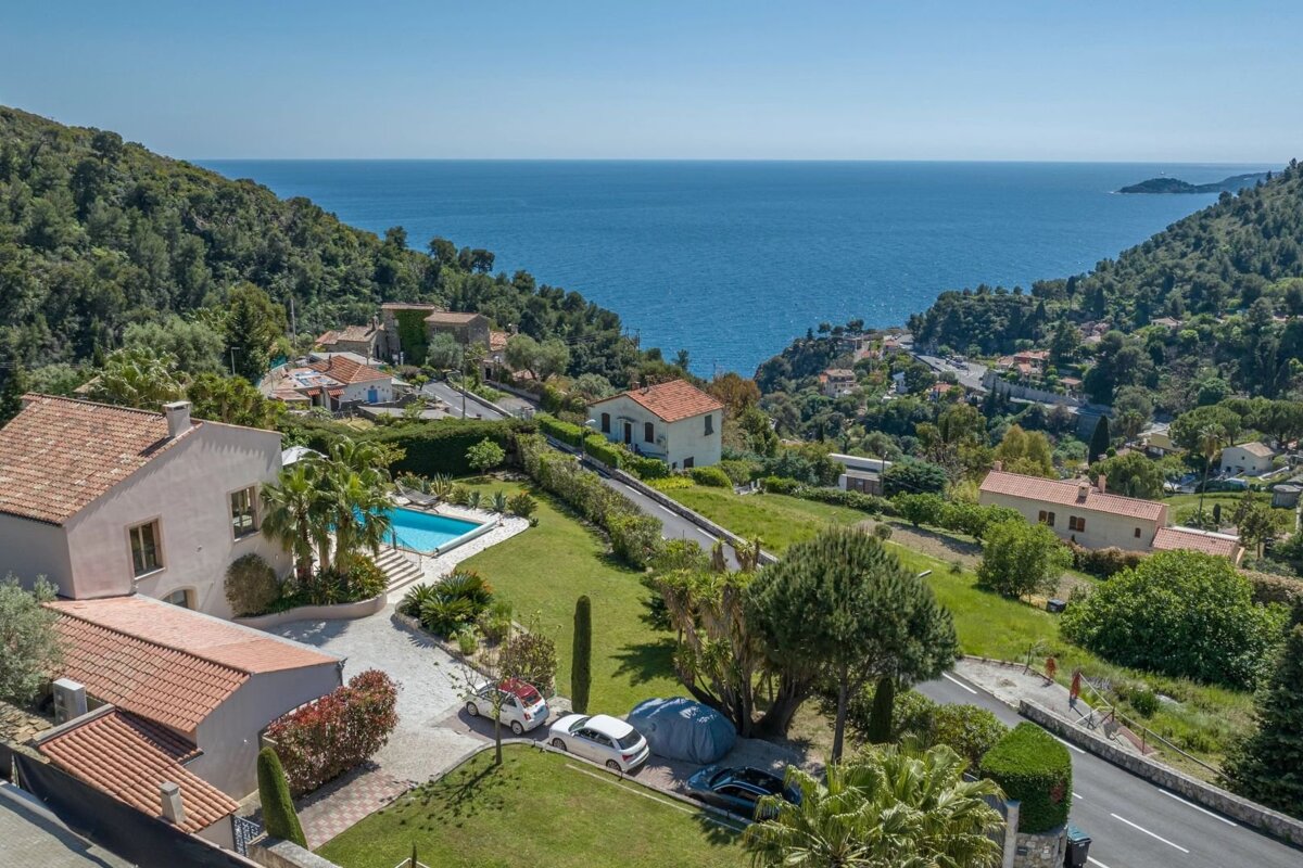 Aerial view of a Mediterranean coast with houses, a pool, lush green hills, and a clear blue sea under a sunny sky.