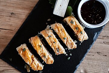 Slices of golden-brown pastry garnished with herbs, served on a black slate platter with a wedge of cheese and dark dipping sauce.
