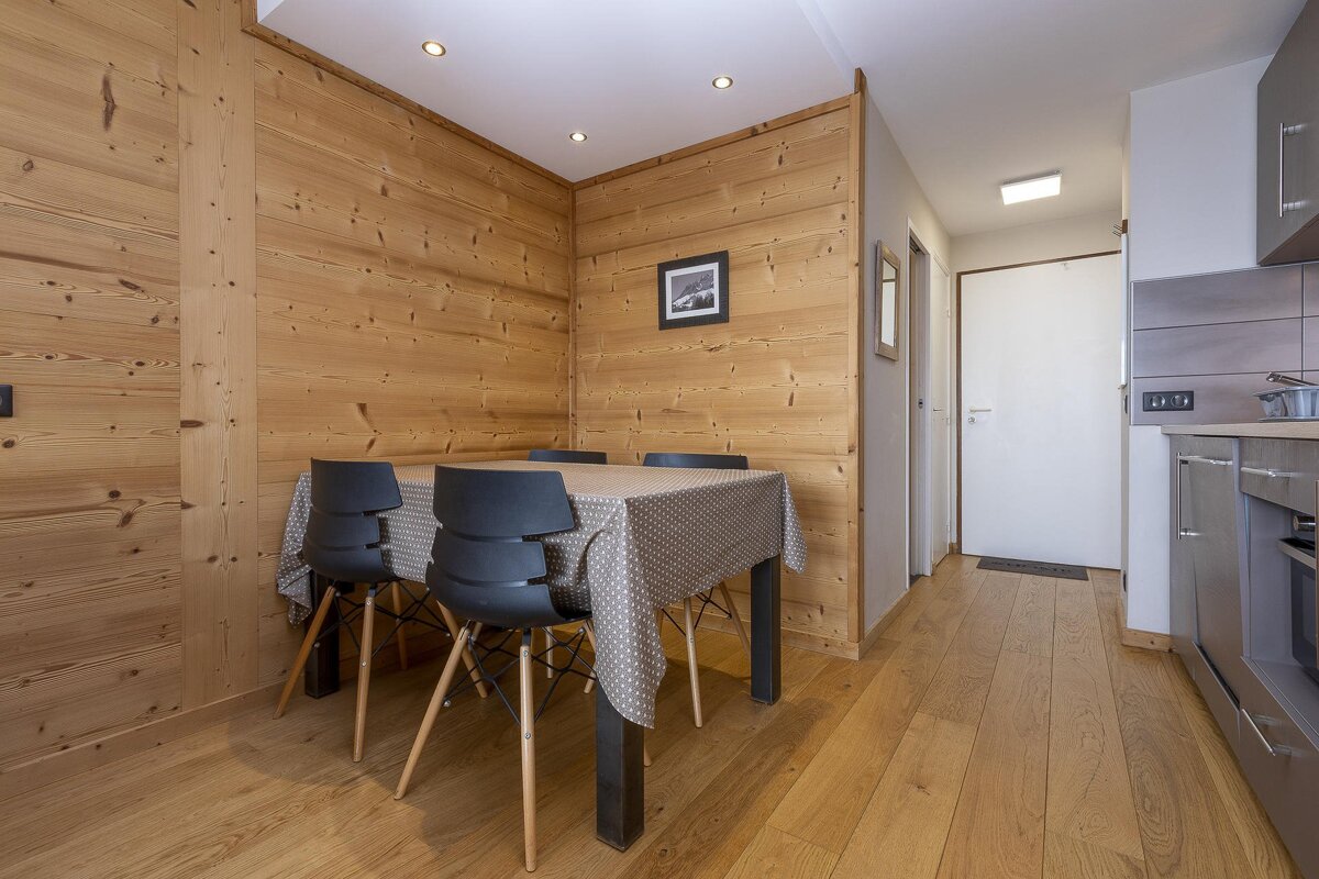 Cozy dining space with wood-paneled walls, a table, black chairs, and a dotted tablecloth. A modern kitchen and hallway are visible.