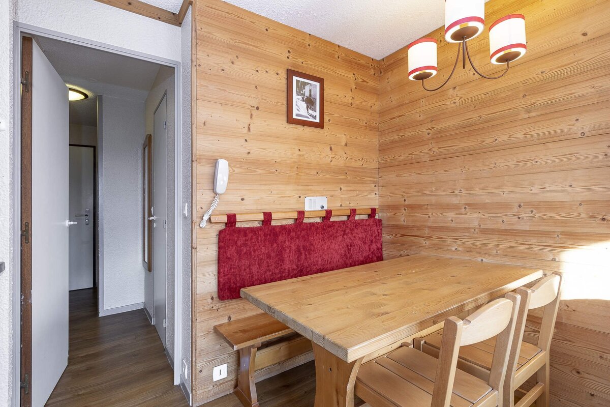 A cozy, rustic dining area with light wood paneling, a wooden table, chairs, and a red upholstered bench. A hallway is visible to the left.