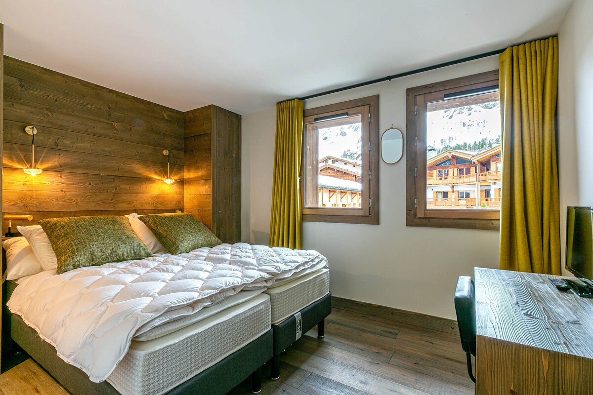 Cozy bedroom with wooden headboard & floor, twin beds, and yellow curtains. Windows show snowy mountain chalets. A desk and TV complete the room.