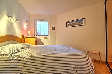 A well-lit, cozy bedroom featuring a striped bed, wooden nightstand with a lamp, window with heart-patterned curtains, and a mountain picture on the wall.