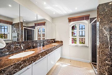 A luxurious bathroom featuring a dark marble double vanity, white cabinets, a large mirror, a shower, and a window with a vibrant outdoor view.