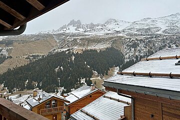 View of a snow-dusted mountain village with wooden chalets and pine forests, set against rugged, snow-capped peaks under a cloudy sky.