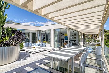 A chic outdoor terrace features a modern lounge, dining area under a pergola, lush greenery, and glass railings with expansive views.