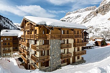A large wooden and stone chalet-style building with balconies, covered in snow, nestled in a snowy mountain resort under a blue sky.