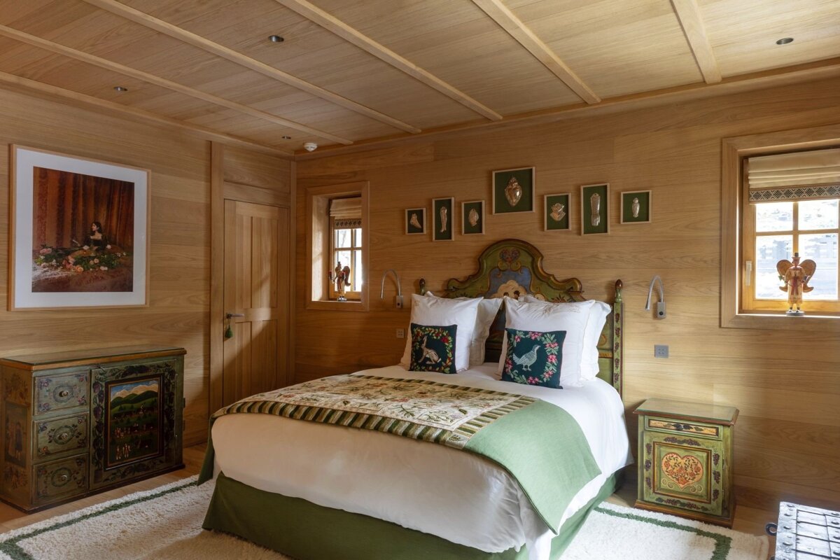 A cozy, wood-paneled bedroom with a decorative green bed, painted folk art furniture, and framed wall art, creating a rustic atmosphere.
