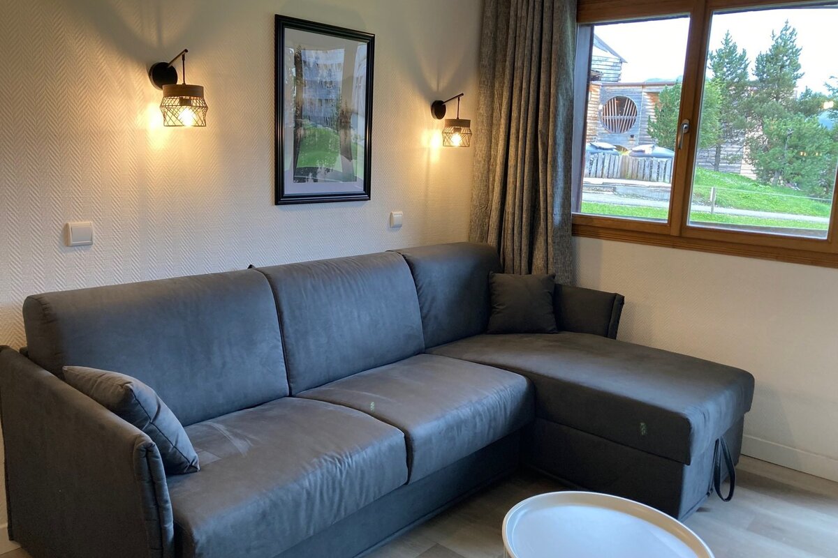 A cozy living room with a large grey sectional sofa, wall sconces, and a framed picture. A window offers a view of green trees and a building.