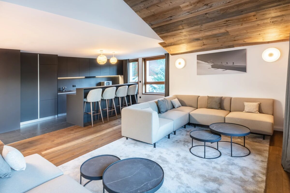 Modern open-plan living room and dark kitchen featuring a large sectional sofa, nested coffee tables, and a wooden ceiling.