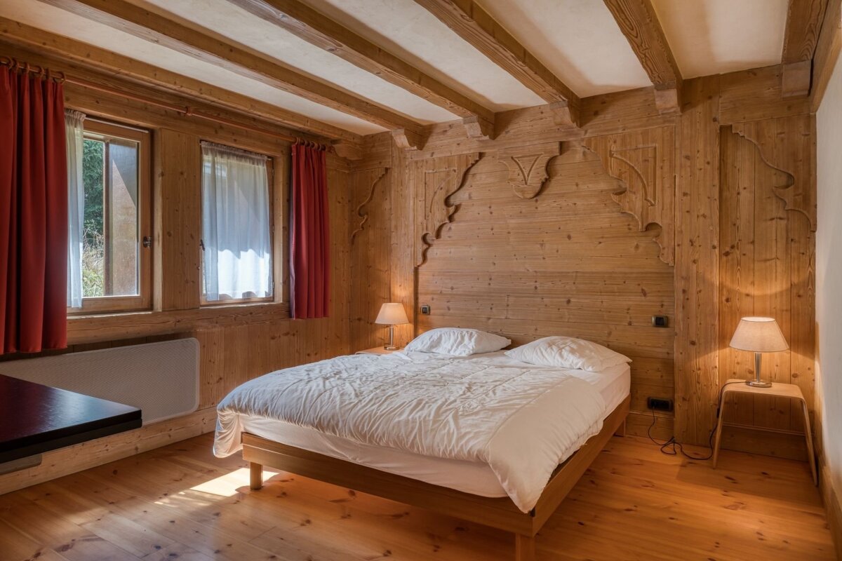 A warm, rustic bedroom with full wood paneling, a bed with white linen, two windows with red curtains, and two bedside lamps.
