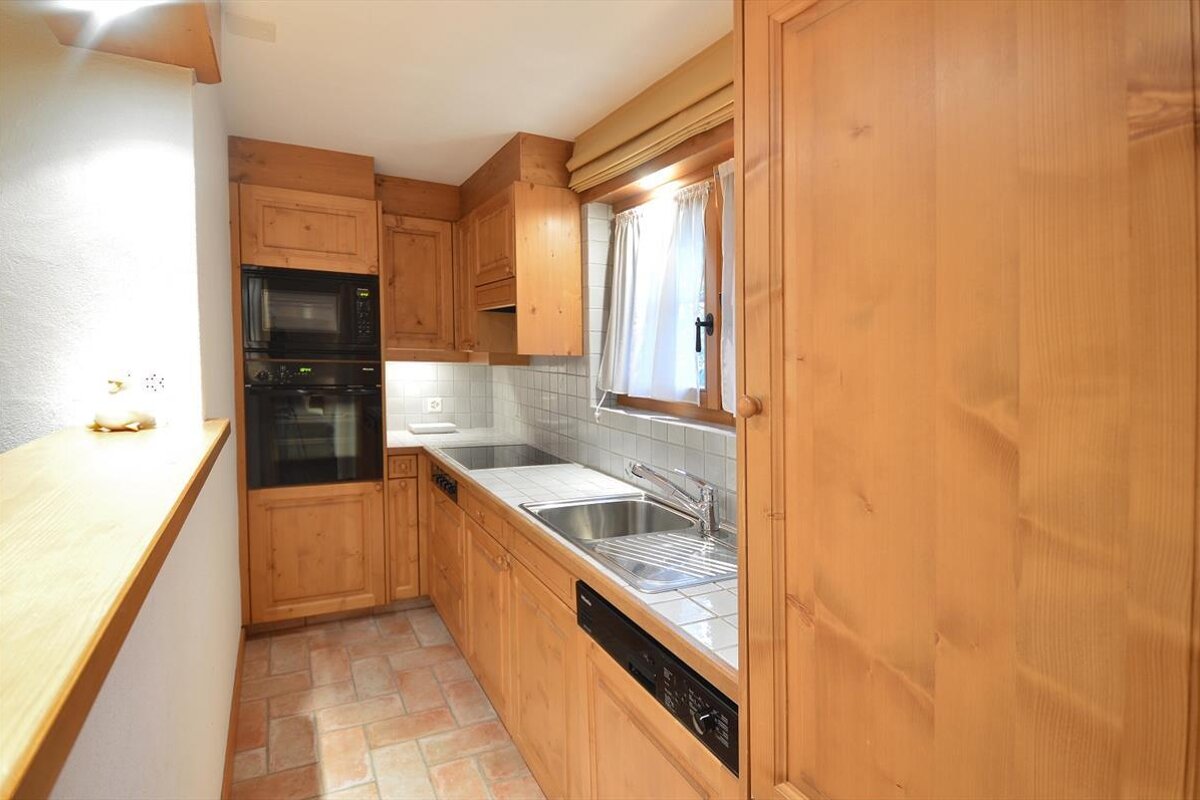 A rustic kitchen with natural wood cabinets, terracotta tiled floor, built-in appliances (oven, microwave), and a window with white curtains.