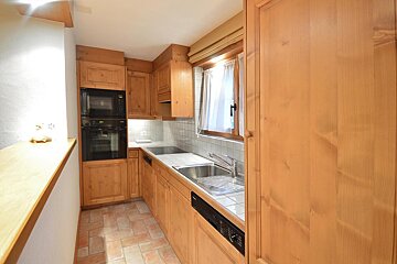 A rustic kitchen with natural wood cabinets, terracotta tiled floor, built-in appliances (oven, microwave), and a window with white curtains.