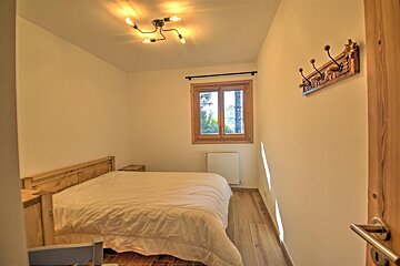 A cozy bedroom features a wooden bed with a white duvet, a window overlooking trees, a unique ceiling light, and a carved wooden coat rack.