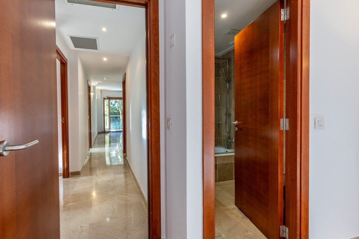 A bright hallway with shiny tiled floors and multiple open brown doors, one revealing a bathroom with a shower and tub.