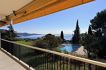Sunny balcony view with yellow awning, overlooking a blue sea, green garden, and a swimming pool.