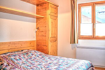 Cozy bedroom with a colorful quilted bed, wooden headboard, shelf, and built-in cabinet. A wooden-framed window with curtains and a heater are visible.