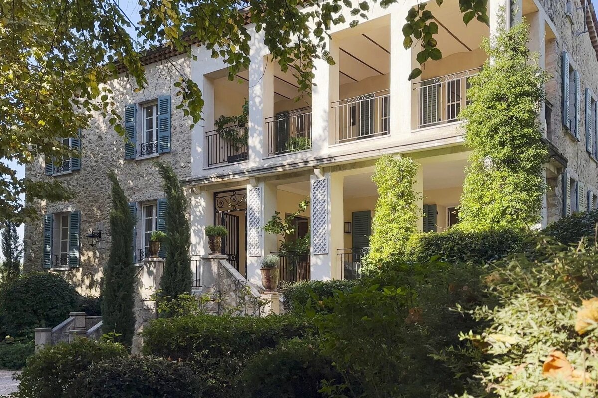 A two-story stone mansion with blue shutters and a white-columned balcony is surrounded by lush green trees and bushes.