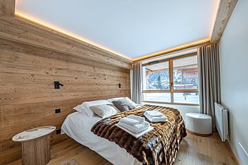 Cozy chalet bedroom with wood-paneled walls, warm lighting, a fur blanket on the bed, and a window looking out at a snowy landscape.