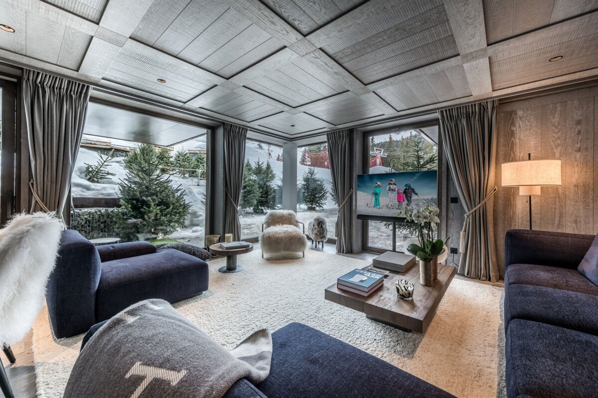 Cozy ski chalet living room with panoramic snowy views, featuring plush sofas, a furry chair, and warm wooden accents throughout.