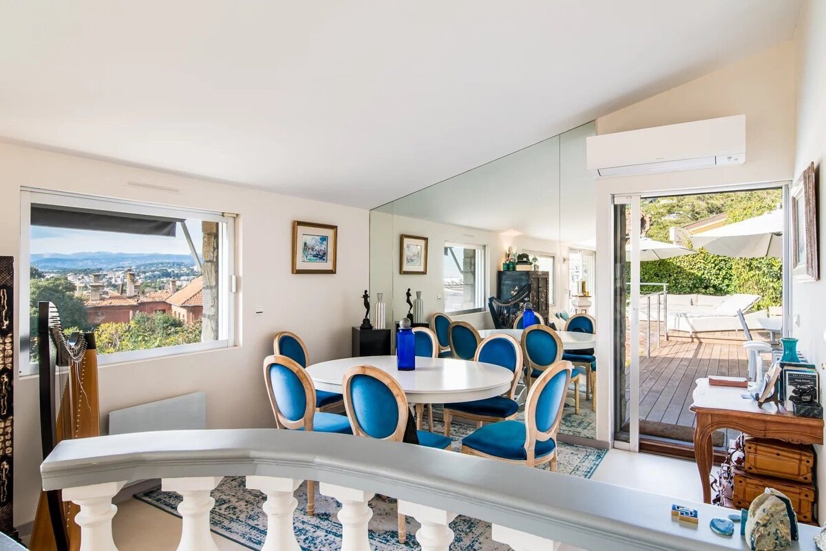 A bright dining room with a white round table, blue velvet chairs, a large window overlooking a city, and access to an outdoor deck.