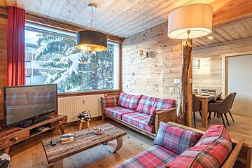 Cozy wooden chalet living room featuring red plaid sofas, rustic furniture, and a scenic view of snowy trees.