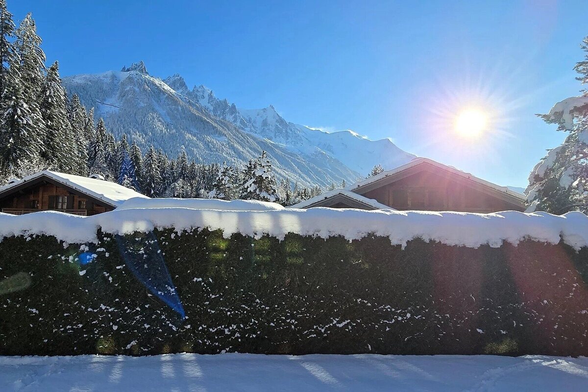 Sun-drenched snowy mountain landscape with wooden chalets, pine trees, and a snow-covered hedge against a clear blue sky.