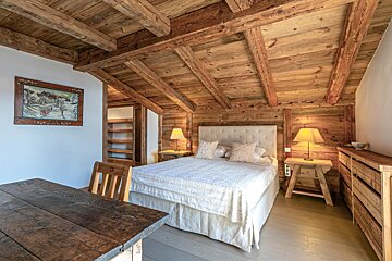 A rustic wooden chalet bedroom features a large bed, exposed wood beams, a distressed table, and a winter painting on the wall.