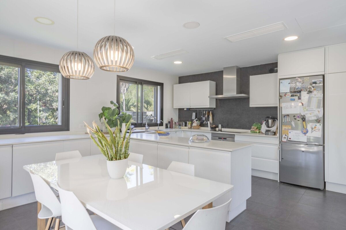 Bright, modern kitchen with white cabinets, dark backsplash, a dining area, large island, stylish pendant lights, and big windows.