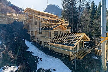 A multi-level chalet under construction with scaffolding on a snowy mountain slope surrounded by pine trees.