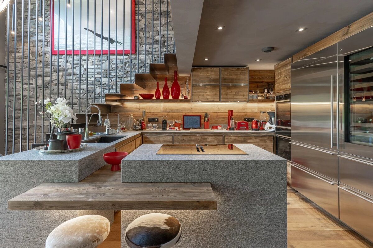 Modern rustic kitchen featuring large stone islands, wooden cabinetry, stainless steel appliances, and red accents against a stone wall.