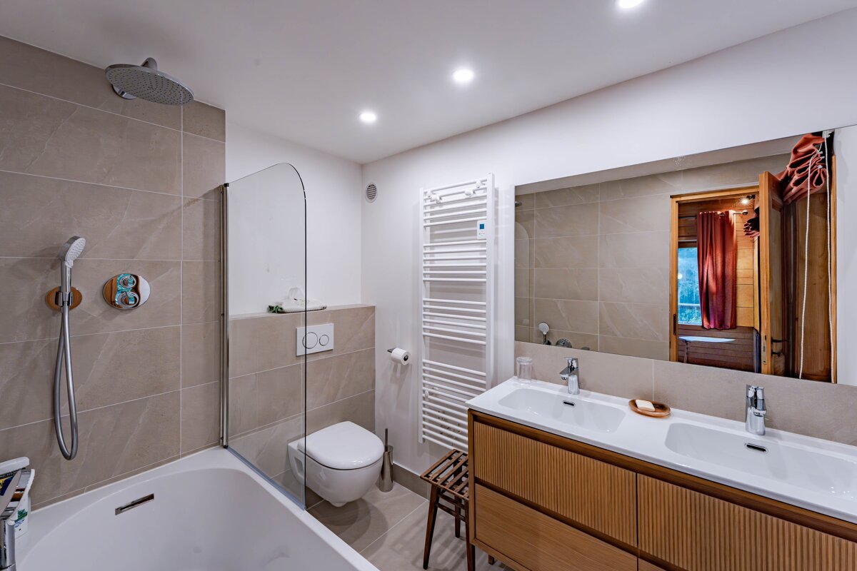 Bright, modern bathroom with a tub/shower, toilet, double wood vanity, large mirror, and towel warmer, featuring neutral tiles.