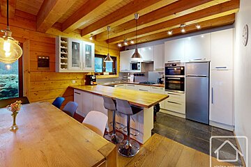 Modern kitchen with wood walls & exposed beams, dining table, and breakfast bar in a cozy chalet. Features white cabinetry and appliances.