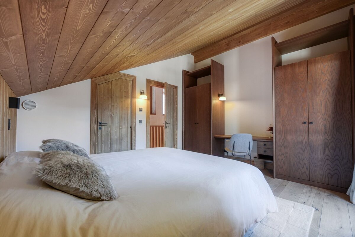 Cozy bedroom with slanted wooden ceiling, large bed, fur pillows, built-in wooden wardrobes, and a desk.