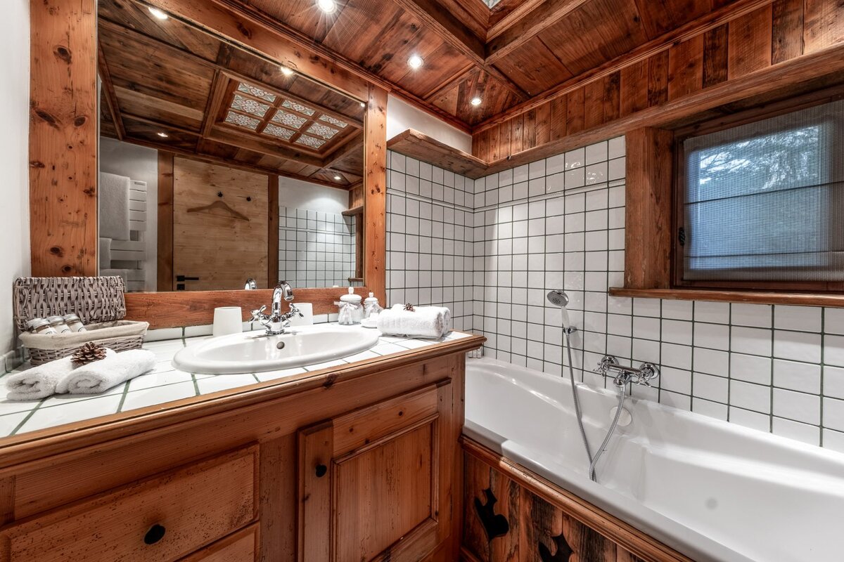 A rustic bathroom features a wood-paneled ceiling, vanity, and mirror, with white tiled walls, a sink, and a clean white bathtub.